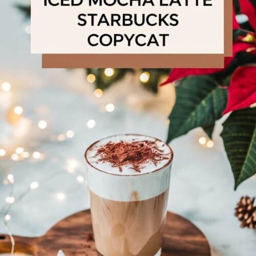 A close-up of a glass of iced mocha latte topped with a layer of whipped cream and dusted with cocoa powder. The drink is presented on a wooden serving board, with soft, blurred holiday lights and a red poinsettia plant in the background. The text overlay reads "Iced Mocha Latte - Starbucks Copycat."